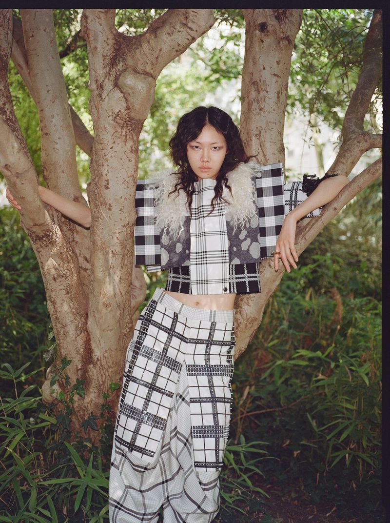 Model Sijia Kang posing between tree branches wearing a black and white checkered outfit in a wooded garden.