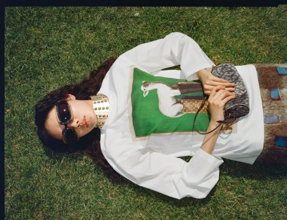 Model Sijia Kang wearing oversized sunglasses, a graphic white shirt, and gold studded choker lying on grass.