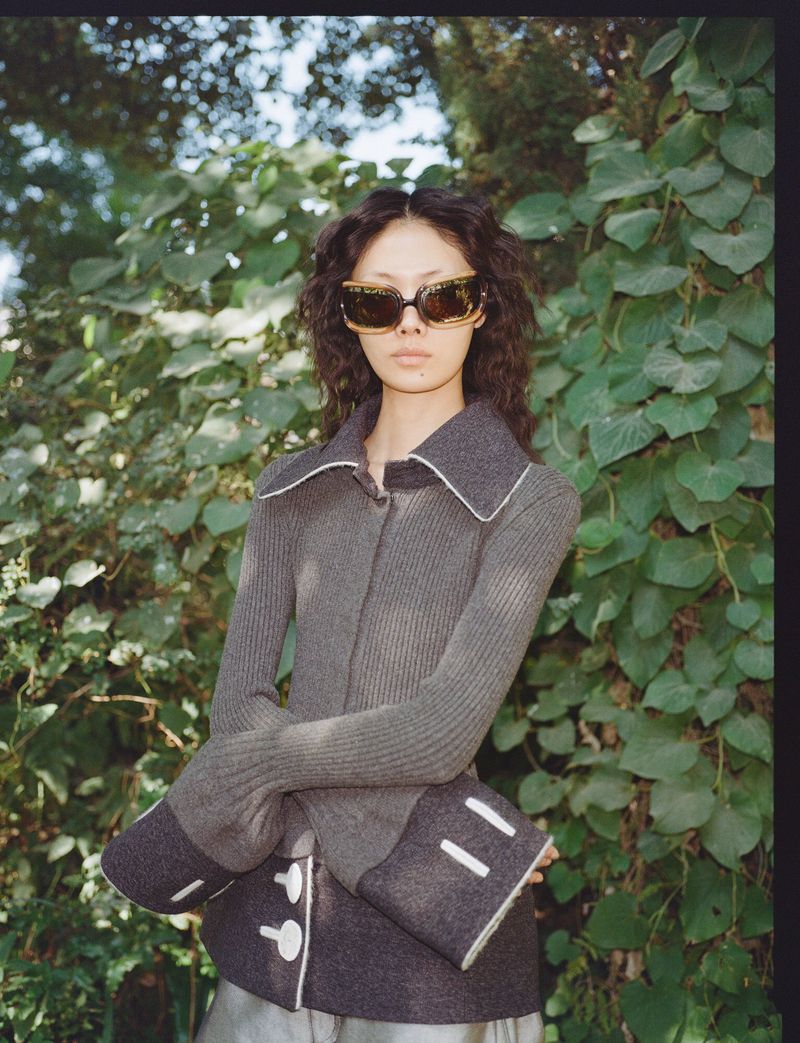 Model Sijia Kang posing in oversized sunglasses and a grey ribbed knit jacket against lush green foliage.