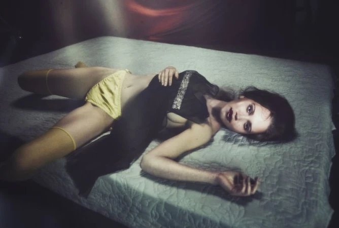 Model Arina Maksimova lies back on a mattress wearing yellow satin underwear, yellow thigh-highs, and a sheer black top.
