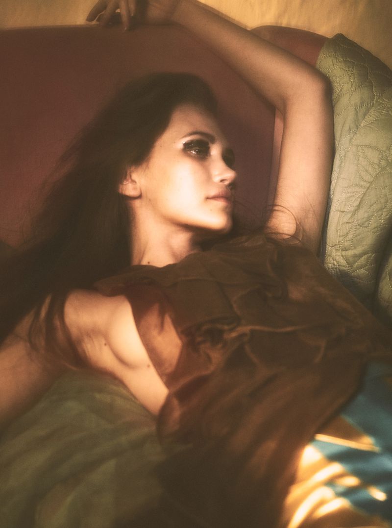 Model Arina Maksimova reclined with arm raised wearing a brown textured draped top under warm soft light.