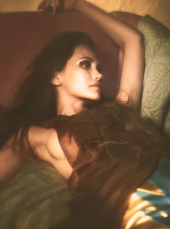 Model Arina Maksimova reclined with arm raised wearing a brown textured draped top under warm soft light.