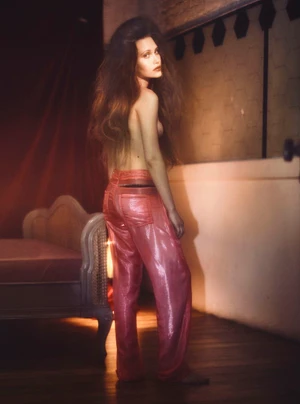 Model Arina Maksimova posing topless in shiny pink trousers looking over her shoulder beside a vintage chaise longue.