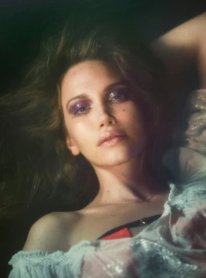 Model Arina Maksimova lying down with shimmery purple eyeshadow and an off-shoulder ruffled top in a moody beauty portrait.