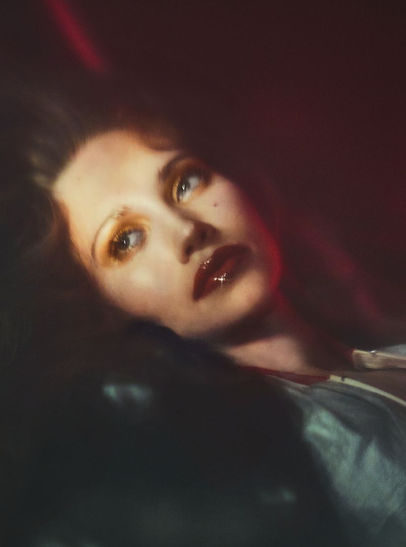 Close-up portrait of model Arina Maksimova featuring glossy dark red lips, yellow eyeshadow, and soft red-tinted ambient lighting.