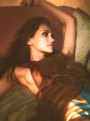 Model Arina Maksimova reclining against cushions with an arm raised in a warm softly lit editorial portrait.