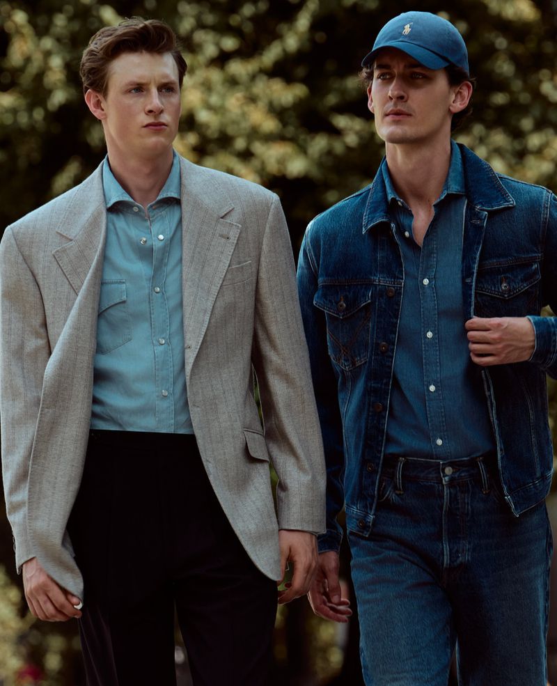 Two men walking outdoors, one in a light grey blazer and blue shirt, the other in a denim jacket, jeans, and a blue baseball cap.