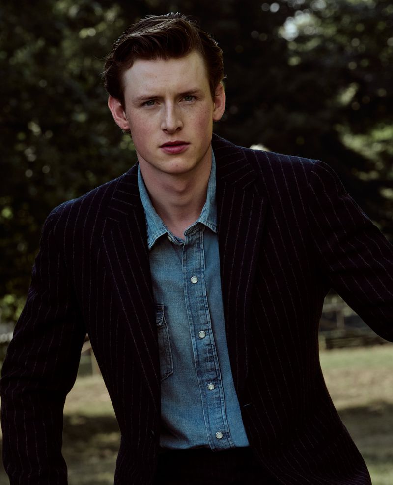 Young man in a dark pinstripe blazer over a light blue denim shirt looking directly forward against an outdoor park background.