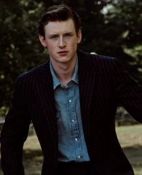 Young man in a dark pinstripe blazer over a light blue denim shirt looking directly forward against an outdoor park background.