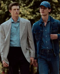 Two men walking together outdoors, one wearing a light grey blazer and blue shirt, the other wearing a denim jacket and blue cap.
