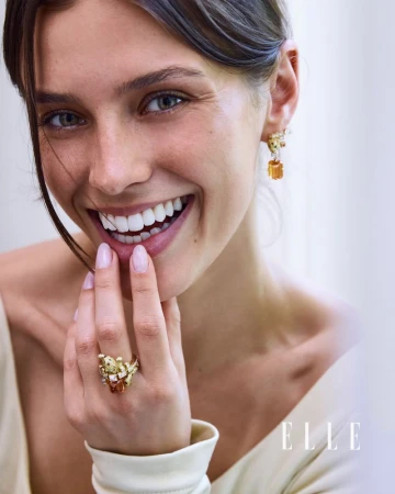 Model Emma Todt smiling while displaying an ornate gold gemstone ring and matching earring against a bright background.