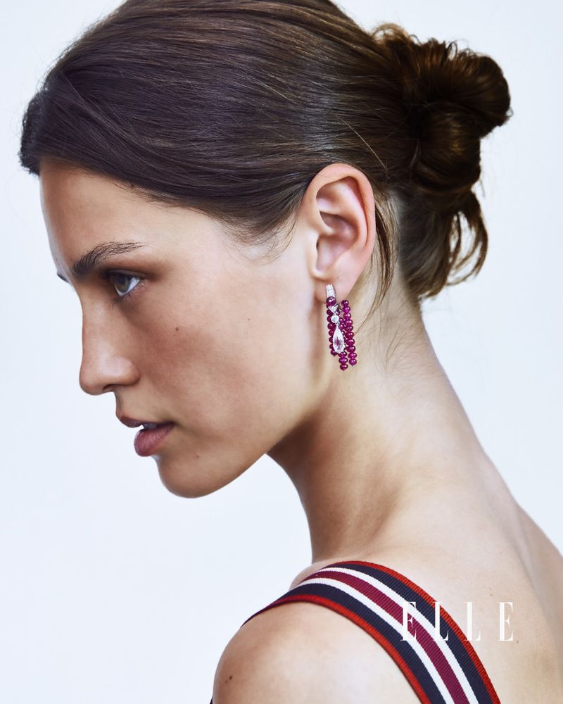 Side profile portrait of a woman with hair in a bun wearing a beaded magenta drop earring against a white studio background.