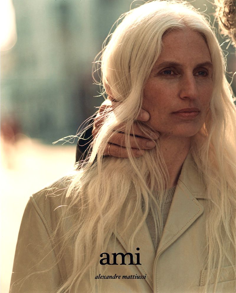 Fashion editorial shot of a fair-skinned adult woman with long blonde hair wearing a tan jacket with a man's hand on her neck.