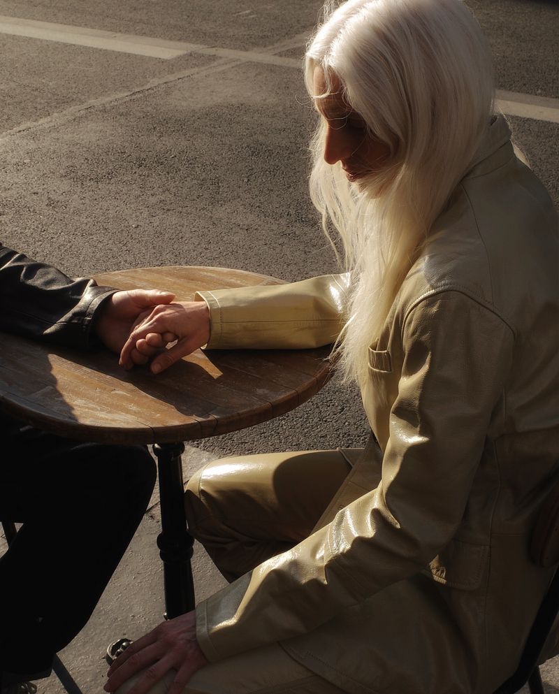Fashion editorial image of a fair-skinned adult woman with long platinum hair holding hands across a round table with a dark-sleeved person.