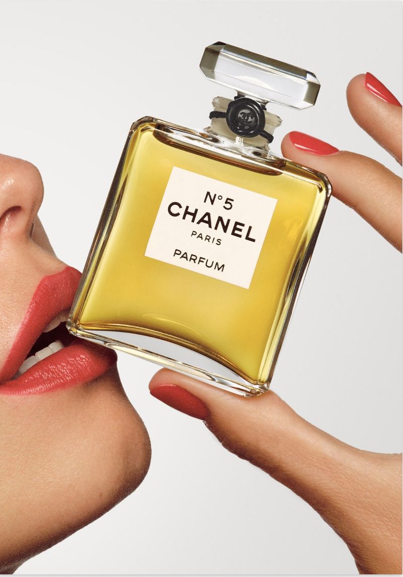 Model with red lipstick and red manicured nails holding a glass Chanel No 5 perfume bottle against a white studio background.