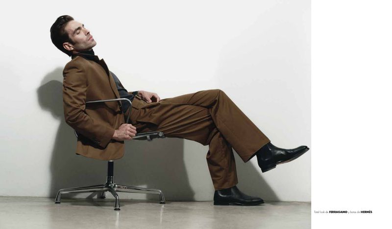 Model Jon Kortajarena reclining in a chrome office chair wearing a brown suit and black boots against a white studio background.