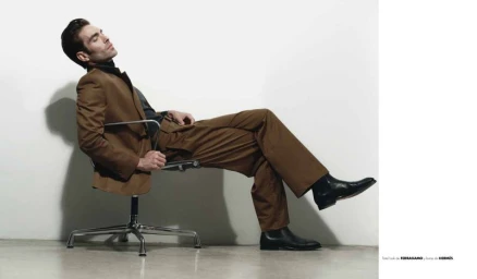 Model Jon Kortajarena reclining in a chrome office chair wearing a brown suit and black boots against a white studio background.