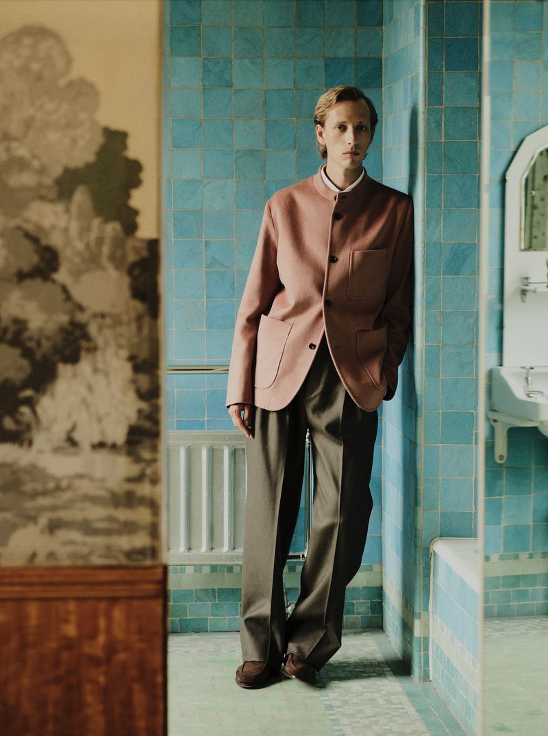 Model Josef Ptacek wearing a dusty rose jacket and pleated wide trousers while leaning against blue tiled walls.