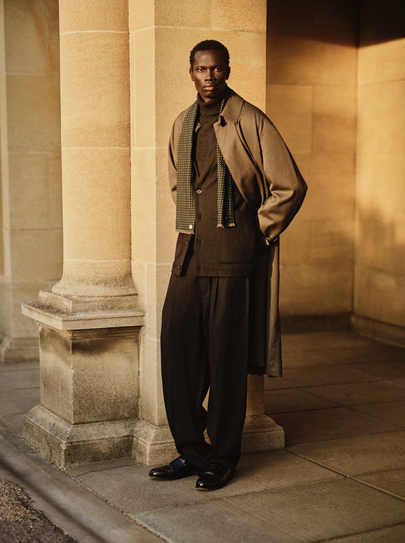Model Makur Mabor in a camel coat, dark tailored suit, and scarf standing beside a sandstone column in warm sunlight.