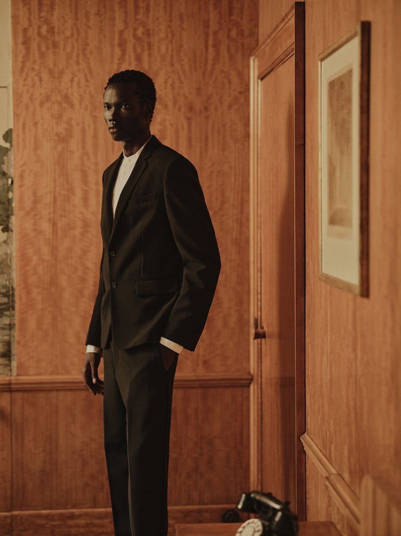 Makur Mabor in a tailored black suit standing inside a warm wood-paneled room with a vintage telephone.