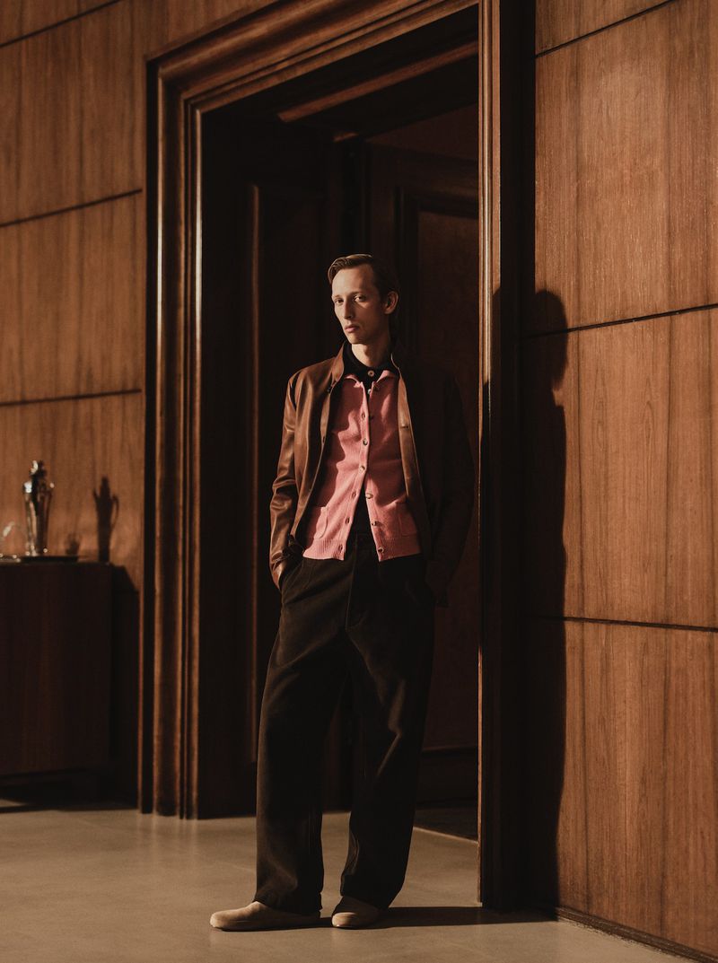 Model Josef Ptacek stands in a wood-paneled doorway wearing a brown leather jacket, pink cardigan, and dark trousers for Telegraph Luxury.