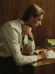 Man in a pinstripe button-down shirt and tie leans over a wooden desk holding a pen with books stacked in the background.