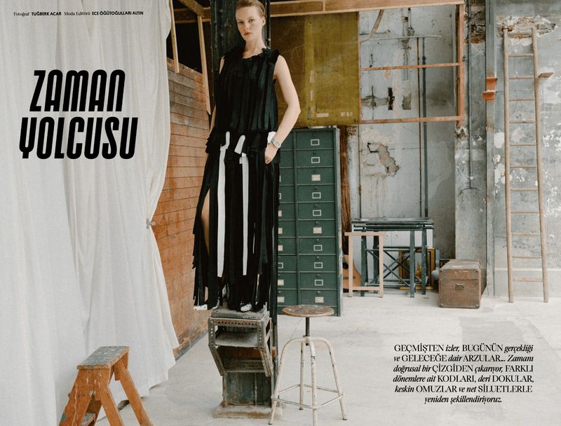 Model Maya Vozhdaeva standing elevated in a black fringed dress in a rustic studio interior with props.
