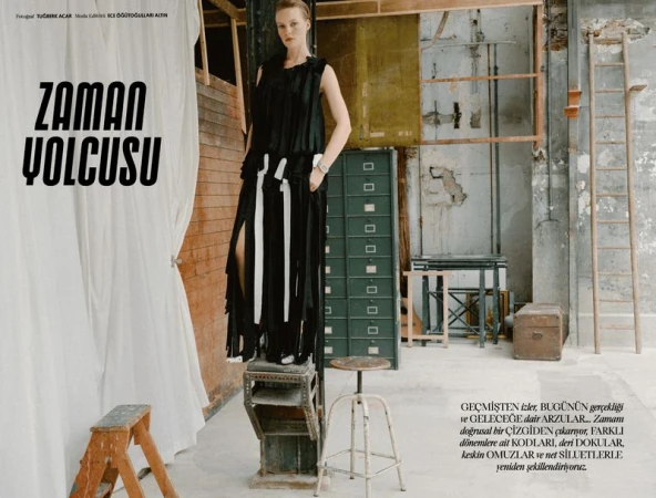 Model Maya Vozhdaeva standing elevated in a black fringed dress in a rustic studio interior with props.