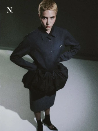 Model with short blonde hair in a black structured puff-waist dress and heels looking up at the camera from a high angle.
