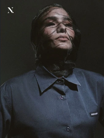 Model wearing a sheer black face veil and a dark navy collared shirt against a dark backdrop for Noir Magazine editorial.