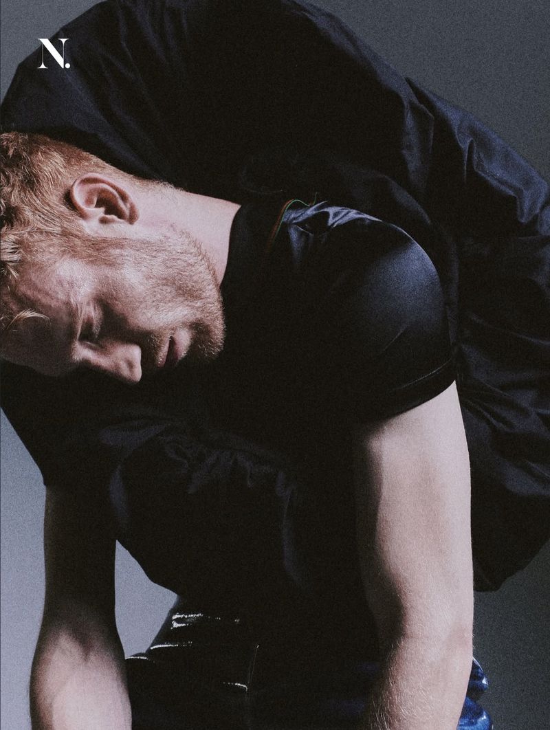 Red-haired man bending forward wearing a black satin top and voluminous black garment against a grey studio backdrop.
