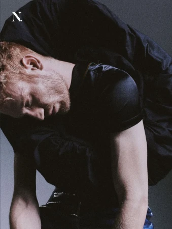 Red-haired man bending forward wearing a black satin top and voluminous black garment against a grey studio backdrop.