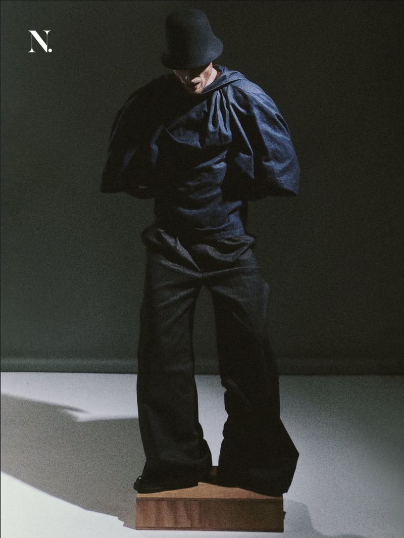 Model in an oversized dark hooded cape top, wide-leg trousers, and a round hat standing on a wooden block in a studio.
