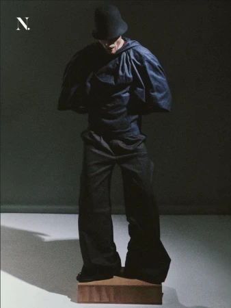 Model in an oversized dark hooded cape top, wide-leg trousers, and a round hat standing on a wooden block in a studio.