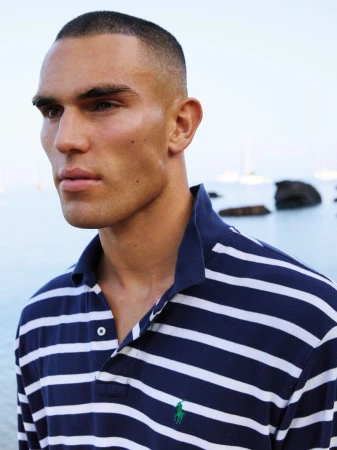 Model Josh McGregor wearing a navy blue and white striped polo shirt outdoors with calm coastal water in the background.