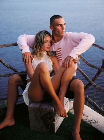 Josh McGregor and a female model sitting intimately on a wooden beam in front of a rustic railing overlooking open water.