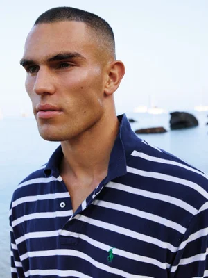 Model Josh McGregor wearing a navy blue and white striped polo shirt in an outdoor coastal setting.
