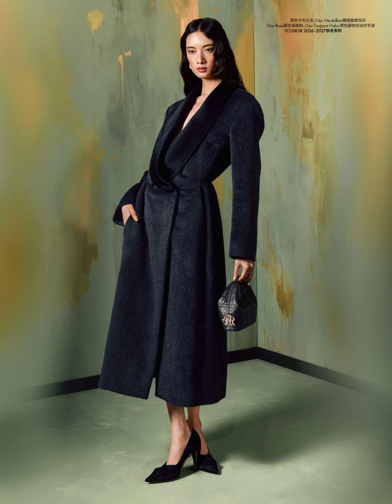 Model Cici Xiang Yejing wearing a tailored long black coat and heels while holding a small handbag against a painterly studio backdrop.