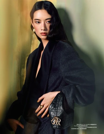 Model Cici Xiang Yejing posing in a charcoal wool coat holding a black quilted handbag against an olive textured backdrop.