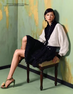 Cici Xiang Yejing poses on a wooden chair in a black dress, cropped white jacket, and green heels against a textured painted wall.