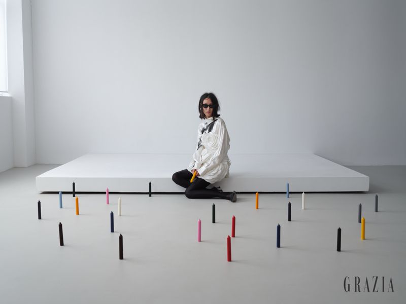 Model Xiru Yang in a white textured blouse, dark sunglasses, and black tights seated by colorful taper candles in a white studio.
