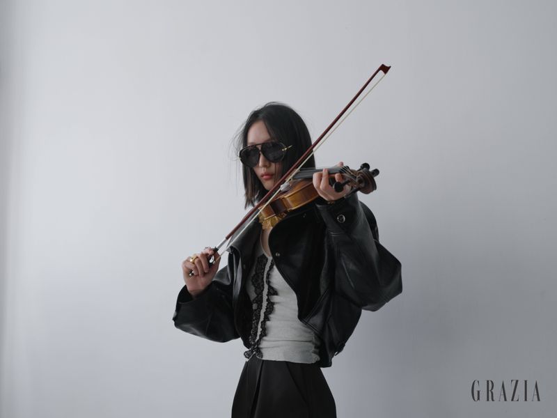 Model wearing dark sunglasses and a cropped black leather jacket playing a violin against a plain studio background.