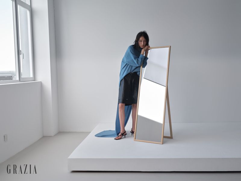 Model Xiru Yang wearing a blue draped top and black shorts leans on a standing floor mirror in a minimalist white studio.