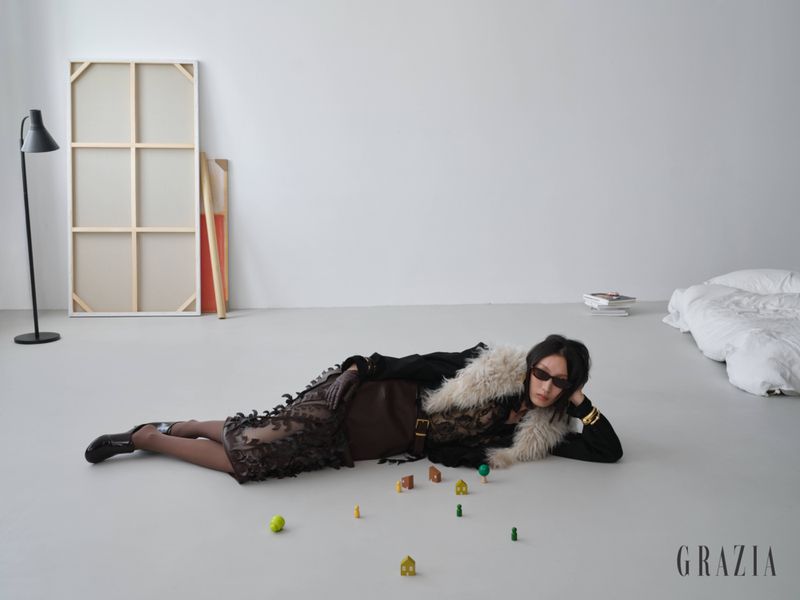 Model Xiru Yang wearing sunglasses and a fur-trimmed dark outfit lying on a studio floor beside miniature houses for Grazia Slovenia.