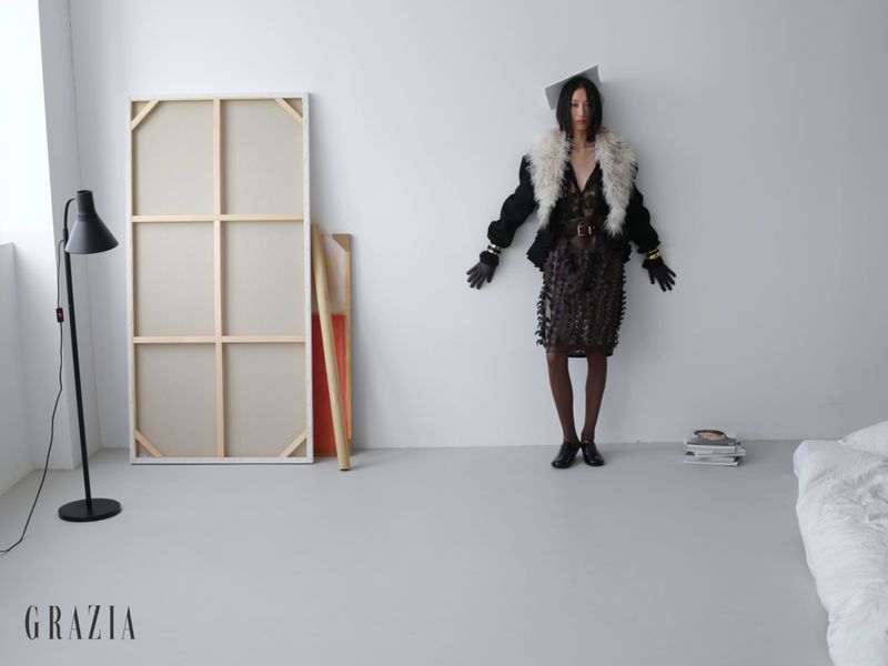 Model Xiru Yang stands against a white studio wall wearing a dark jacket with a white shearling collar, dark lace skirt, and gloves.