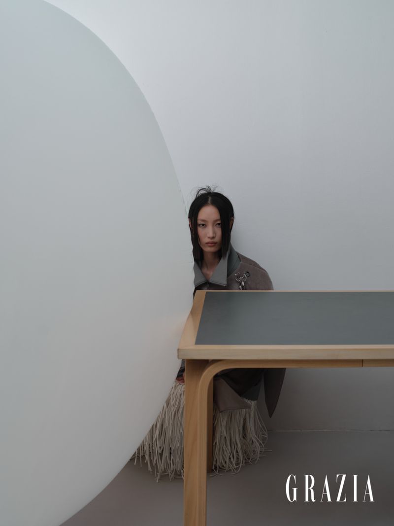 An East Asian woman seated behind a wooden desk and large curved white partition in a minimalist interior setting.