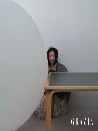 An East Asian woman seated behind a wooden desk and large curved white partition in a minimalist interior setting.