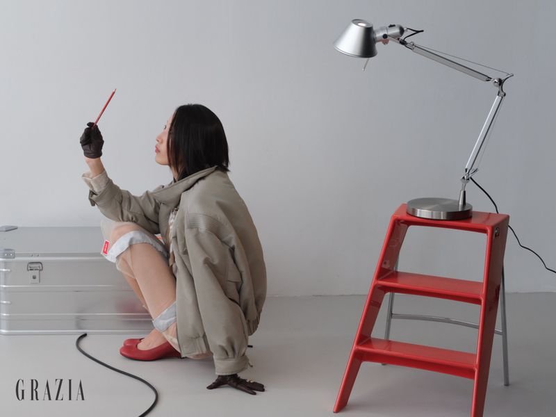 Model Xiru Yang crouching in a taupe bomber jacket and red ballet flats next to a red step stool with a silver desk lamp in studio.