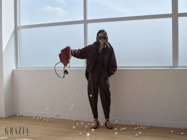 Model Xiru Yang wearing a dark brown leather suit and sunglasses, spilling white balls from a handbag in a bright loft space.