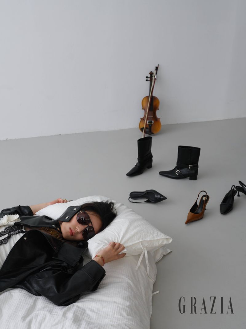 Model wearing a black leather jacket and sunglasses lies on a white pillow near scattered shoes and a violin on a grey studio floor.